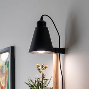 May include: A wall-mounted lamp featuring a matte black conical shade and a light-toned wooden cylindrical support arm. The lamp emits a warm white light directed downward onto a green succulent plant with small yellow flowers positioned below.