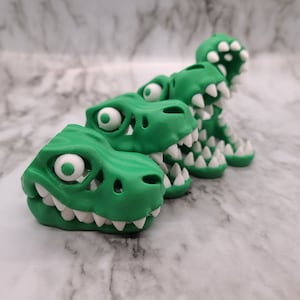 May include: Three green dinosaur head figurines with white teeth and eyes. The figurines are arranged in a row, with the largest figurine in the back and the smallest figurine in the front.