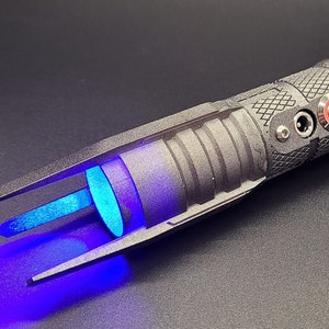 May include: A black lightsaber with a blue blade and a red button. The lightsaber has a textured surface and a silver accent on the handle.