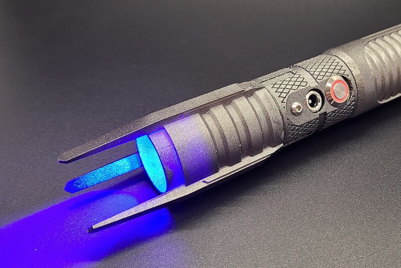 May include: A black lightsaber with a blue blade and a red button. The lightsaber has a textured surface and a silver accent on the handle.