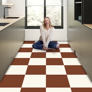 Op de afbeelding: Een vrouw zit op een bruin-witte geruite vloer in een keuken. De keuken heeft witte muren, een raam met jaloezieën en donkere kasten.