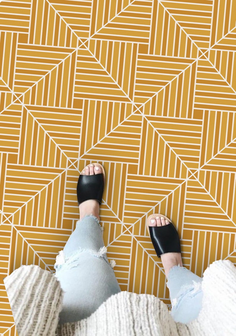 May include: A person wearing blue jeans and a white sweater stands on a floor with a geometric pattern of white lines on a mustard yellow background.