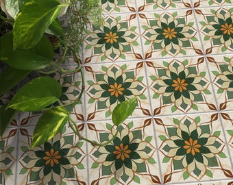 May include: Close-up of patterned tiles with a floral design in shades of green, orange, and brown. The tiles are arranged in a grid pattern, with a cream-colored background. Green leafy plants are in the upper left corner.