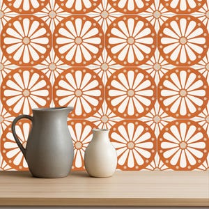 Op de afbeelding: Een stilleven met twee keramische vazen en een kan op een houten oppervlak. De achtergrond is een herhalend patroon van oranje en witte bloementegels. De kan is grijs en de vaas is wit.