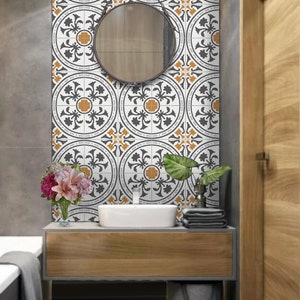 May include: A white bathroom vanity with a gray countertop and a round mirror. The vanity has a wooden cabinet with a gray sink and a gray wastebasket. The wall behind the vanity is tiled with white and black patterned tiles with orange accents.