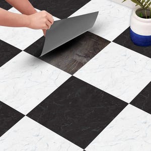 May include: A person installing black and white chequered floor tiles. The tiles have a marble effect. The person is peeling off the backing of the tile. A potted plant with green leaves is in the background.