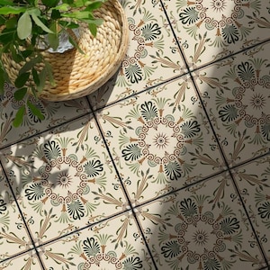 Op de afbeelding: Vierkante keramische vloertegels met een crèmekleurige achtergrond en een herhalend symmetrisch medaillonpatroon in olijfgroen, saliegroen en terracotta. Een gevlochten rieten mand met een groene bladplant staat op de betegelde ondergrond.
