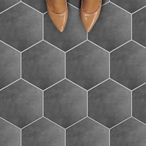 May include: Overhead shot of a person's feet wearing tan pointed-toe heels, standing on a gray hexagonal tile floor. The tiles are outlined with white grout, creating a geometric pattern. The person is wearing dark blue jeans.