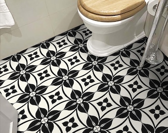 May include: A bathroom floor featuring square tiles with a repeating black and white floral geometric pattern. A white toilet with a light brown wooden seat is positioned in the corner, next to a chrome toilet paper holder and a white tiled wall.