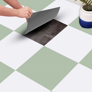 Puede incluir: Una persona está despegando una lámina de baldosa de suelo autoadhesiva gris para revelar una baldosa de imitación de madera marrón debajo. El suelo es un tablero de ajedrez de baldosas blancas y verde claro.