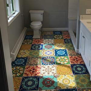 May include: A bathroom floor covered in square tiles featuring a variety of colorful, intricate mandala-style patterns in shades of blue, yellow, green, orange, and red. The floor is situated in a room with gray walls, a white toilet, and a white vanity.