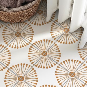 Op de afbeelding: Een witte tegelvloer met een herhalend patroon van goudbruine, paardebloemachtige cirkelvormige motieven. Elk motief bestaat uit een centrale cirkel met uitstralende lijnen die eindigen in kleine stippen, gerangschikt in een raster met grijze voegen.