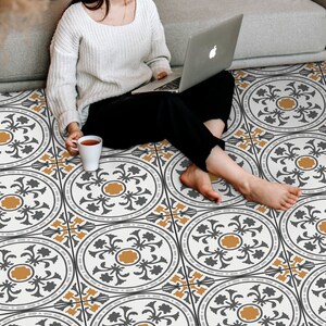 May include: A woman sits on a patterned floor with a laptop on her lap. The floor is covered in white tiles with a black and gold geometric design.