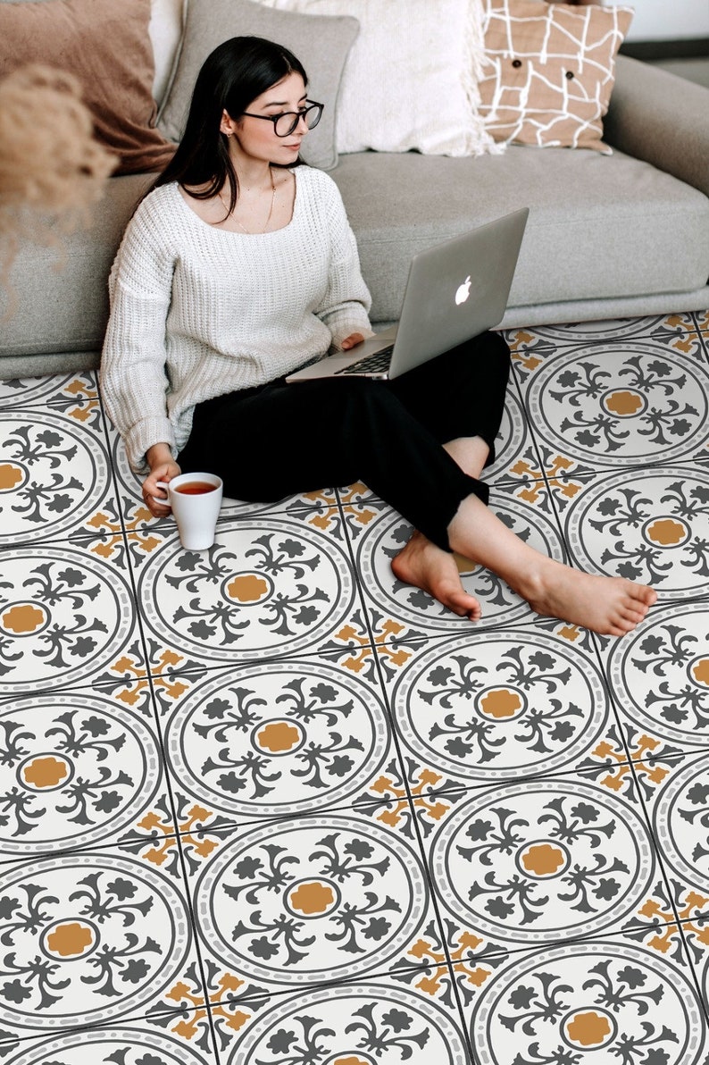 May include: A woman sits on a patterned floor with a laptop on her lap. The floor is covered in white tiles with a black and gold geometric design.