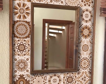 Puede incluir: Espejo rectangular con marco de madera. El marco está decorado con baldosas florales marrones y blancas. El espejo refleja una habitación con puerta y escalera de madera. El espejo está colgado en una pared blanca.