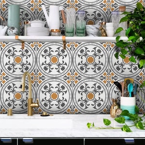 May include: A kitchen backsplash with a white background and a repeating pattern of gray and gold floral designs. The backsplash is behind a white countertop with a gold faucet and a black cabinet below.