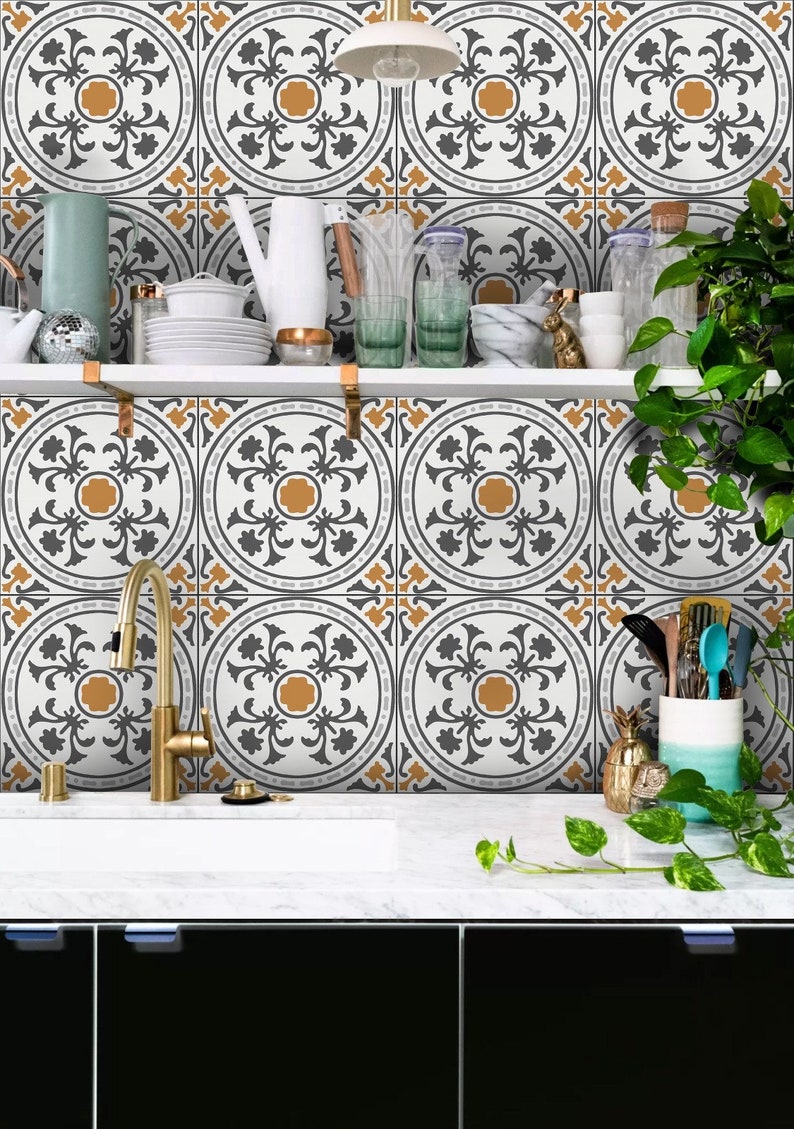 May include: A kitchen backsplash with a white background and a repeating pattern of gray and gold floral designs. The backsplash is behind a white countertop with a gold faucet and a black cabinet below.