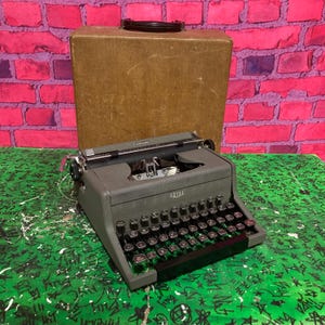 Puede incluir: Una máquina de escribir Royal gris vintage con teclas negras y un carro cromado. La máquina de escribir está sobre una superficie verde con graffiti, frente a una caja marrón y una pared de ladrillos rosas.