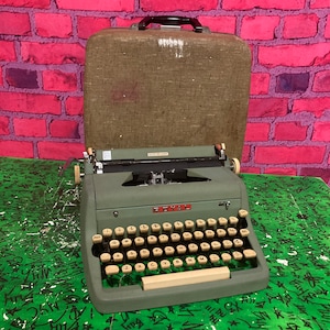 以下が含まれることがあります： ベージュのケースに入った緑色のロイヤル「ポータブル・デラックス」タイプライター。タイプライターには黒いキャリッジリターンレバーと、黒いキーが付いたベージュのキーボードが付いています。
