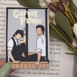 May include: A rectangular sticker with the text "COFFEE PRINCE" in a light yellow font. The sticker features an illustration of two figures in a window frame. One figure is leaning, and the other is sitting.