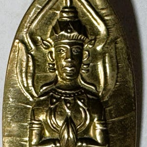 May include: A gold-colored, elongated pendant featuring a detailed relief of a figure with hands clasped in prayer. The figure is adorned with ornate details and surrounded by stylized elements. The pendant has a black base.