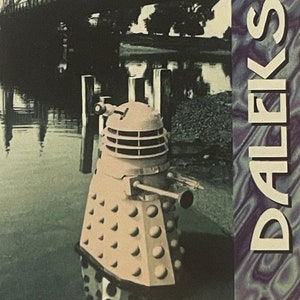 May include: A vintage-style postcard featuring a Dalek from Doctor Who. The image shows a Dalek near a bridge over water. The word "DALEK!" is printed vertically on the right side of the card. The Doctor Who logo is in the upper right corner.