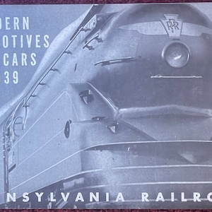 May include: A black and white photo of a Pennsylvania Railroad train engine. The engine is a streamlined locomotive with the text "Pennsylvania Railroad" and "PRR" on the side. The text "MODERN LOCOMOTIVES AND CARS 1939" is printed on the cover of the brochure.