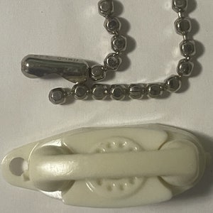 May include: An off-white plastic pulley with a metal ball chain and a metal clasp. The pulley has a curved design with a hole on one end. The metal clasp has the text "B.F. CONN" engraved on it.