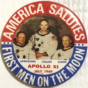 May include: A blue and red round button with the text "America Salutes First Men on the Moon Apollo XI July 1969". The button features a photo of the three Apollo 11 astronauts: Neil Armstrong, Buzz Aldrin, and Michael Collins.