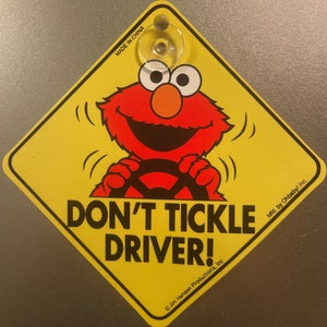 May include: A yellow diamond-shaped car sign with a red Sesame Street character, Elmo, driving a car. The sign reads "Don't Tickle Driver!" with the text "Made in China" and "Mfd. by Ohbaby! Inc." at the top and bottom of the sign. The sign is made by Jim Henson Productions, Inc.