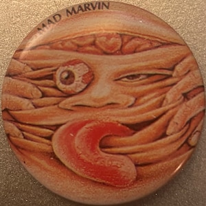 May include: Round button with a surreal illustration of a face with an exposed eye, a red tongue, and the text "MAD MARVIN" at the top. The artwork features a fleshy, textured appearance with a light brown and red color palette.