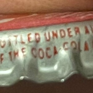 Op de afbeelding: Een zilveren flessen dop met een rode bovenkant. Op de dop staat de tekst "Bottled Under Authority of The Coca-Cola Co.".