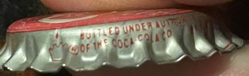 Op de afbeelding: Een zilveren flessen dop met een rode bovenkant. Op de dop staat de tekst "Bottled Under Authority of The Coca-Cola Co.".