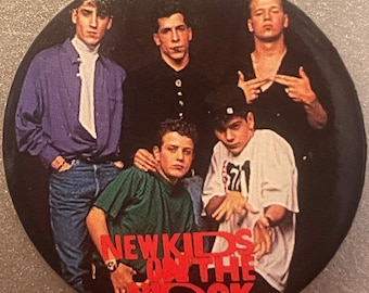 May include: A black and white button featuring a photo of the band New Kids on the Block. The band members are wearing casual clothing and are posing for a photo. The band's name is written in red letters on the button.