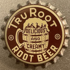 May include: A brown and white bottle cap with the text "Tru Root Delicious and Creamy Root Beer" and a registered trademark symbol on each side. The center of the cap features a cartoon image of a mug filled with root beer.
