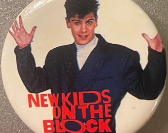May include: A white button with a red and black graphic of a man in a blue suit with the text "NEW KIDS ON THE BLOCK".