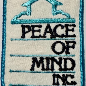 May include: A rectangular embroidered patch with a white background and a teal border. The design features a teal outline of a house roof above the black embroidered text PEACE OF MIND INC. separated by horizontal teal lines on a white fabric base.