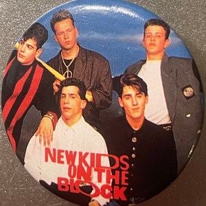 May include: A round button with a picture of five young men. The men are wearing casual clothing and are standing in front of a blue background. The text "NEW KIDS ON THE BLOCK" is printed in red letters on the button.