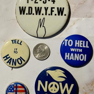 May include: Six vintage peace sign buttons. One button is white with black text that reads "1-2-3-4 W.D.W.Y.F.W." and a peace sign. Another button is white with blue text that reads "Tell it to Hanoi". Another button is blue with white text that reads "To Hell with Hanoi". Another button is blue with white text that reads "Now" and two white doves with olive branches. Another button is blue with a red, white, and blue American flag inside a peace sign. The last button is a silver US quarter.