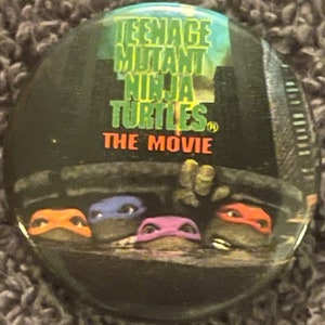 May include: A Teenage Mutant Ninja Turtles button featuring the four turtles in a sewer scene. The button has the movie title "Teenage Mutant Ninja Turtles The Movie" printed on it.