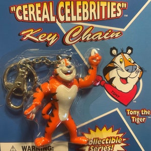 May include: A plastic key chain featuring Tony the Tiger from Frosted Flakes cereal. The keychain is part of a "Cereal Celebrities" collectible series. The image includes a warning about choking hazards for children under 3 years old.