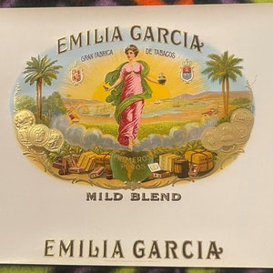 Puede incluir: Una etiqueta de caja de cigarros vintage con un dise&ntilde;o dorado y blanco. La etiqueta presenta a una mujer con un vestido rosa de pie frente a un paisaje con palmeras. El texto en la etiqueta dice "Emilia Garcia Gran Fabrica de Tabacos Mild Blend Emilia Garcia".
