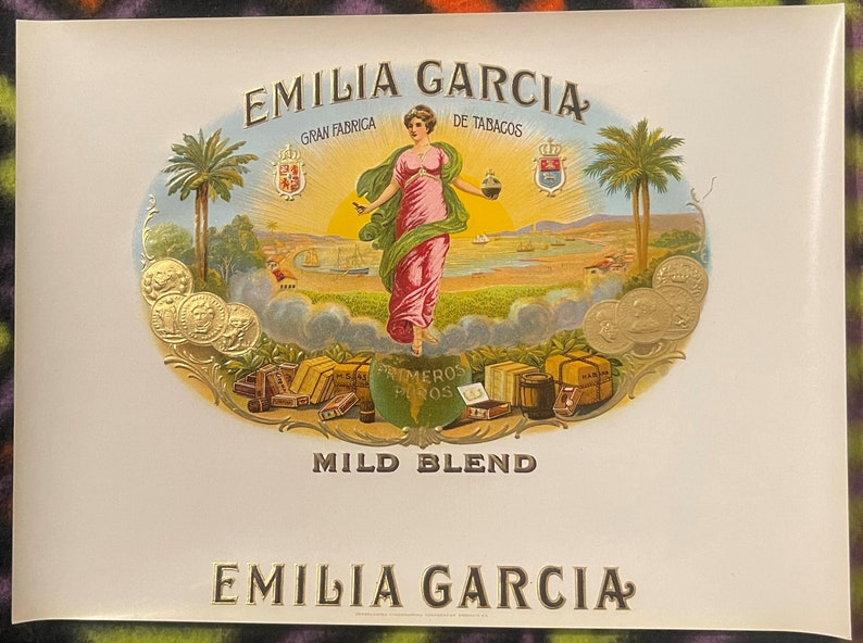 Puede incluir: Una etiqueta de caja de cigarros vintage con un dise&ntilde;o dorado y blanco. La etiqueta presenta a una mujer con un vestido rosa de pie frente a un paisaje con palmeras. El texto en la etiqueta dice "Emilia Garcia Gran Fabrica de Tabacos Mild Blend Emilia Garcia".