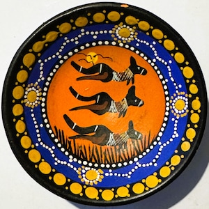 May include: A round, hand-painted ceramic bowl with a black rim and a vibrant orange center. The bowl features a blue and white dotted border, with yellow sun-like designs. The center depicts three stylized animal figures in brown and black.