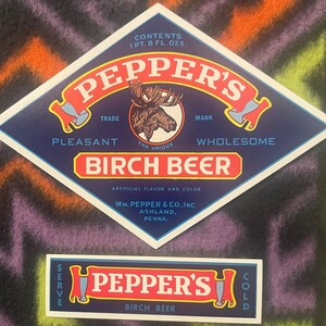 May include: A vintage label for Pepper's Birch Beer, a trademarked beverage. The label features a blue diamond shape with a moose illustration and the text "Pepper's", "Trade Mark", "Pleasant", "The Unique", "Wholesome", and "Birch Beer". The label also includes the company name and location: "Wm. Pepper & Co., Inc., Ashland, Penna.".