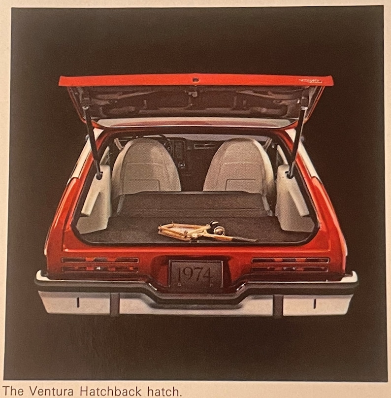 May include: A red 1974 Ventura Hatchback with the boot open, showing the interior. The car has white seats and a black cargo area. A fishing rod is visible in the cargo area. The text "1974" is on the number plate.
