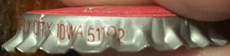 Op de afbeelding: Een zilveren flessen dop met een rode bovenkant. Op de dop staat de tekst "City, Iowa 51102" gedrukt.