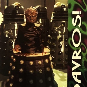 May include: A close-up of a Dalek from the Doctor Who television series. The Dalek is silver and black with a dome-shaped top. The Dalek is surrounded by a green and purple background. The text "DAVROS!" is in the upper right corner of the image.