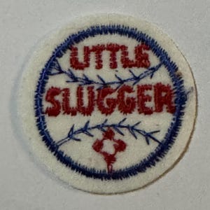 May include: A circular embroidered patch with a white background and a blue stitched border. The text LITTLE SLUGGER is embroidered in red. The design includes blue stitching resembling baseball seams and a small red diamond shape at the bottom.