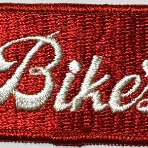 May include: Embroidered red patch with the word "Bike's" in white script. The patch has a red border and a textured appearance. The design is likely for a bicycle-related product or apparel.